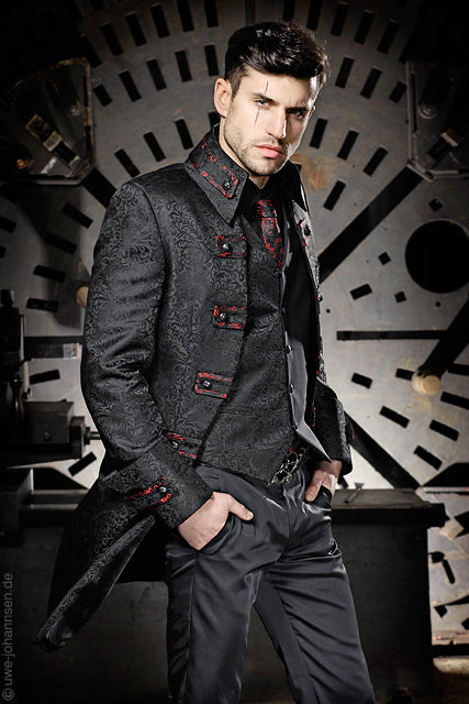 A man wearing a dark patterned double-breasted coat with red button accents, a matching dark vest, and dark trousers, posed in front of a large industrial circular metal structure.