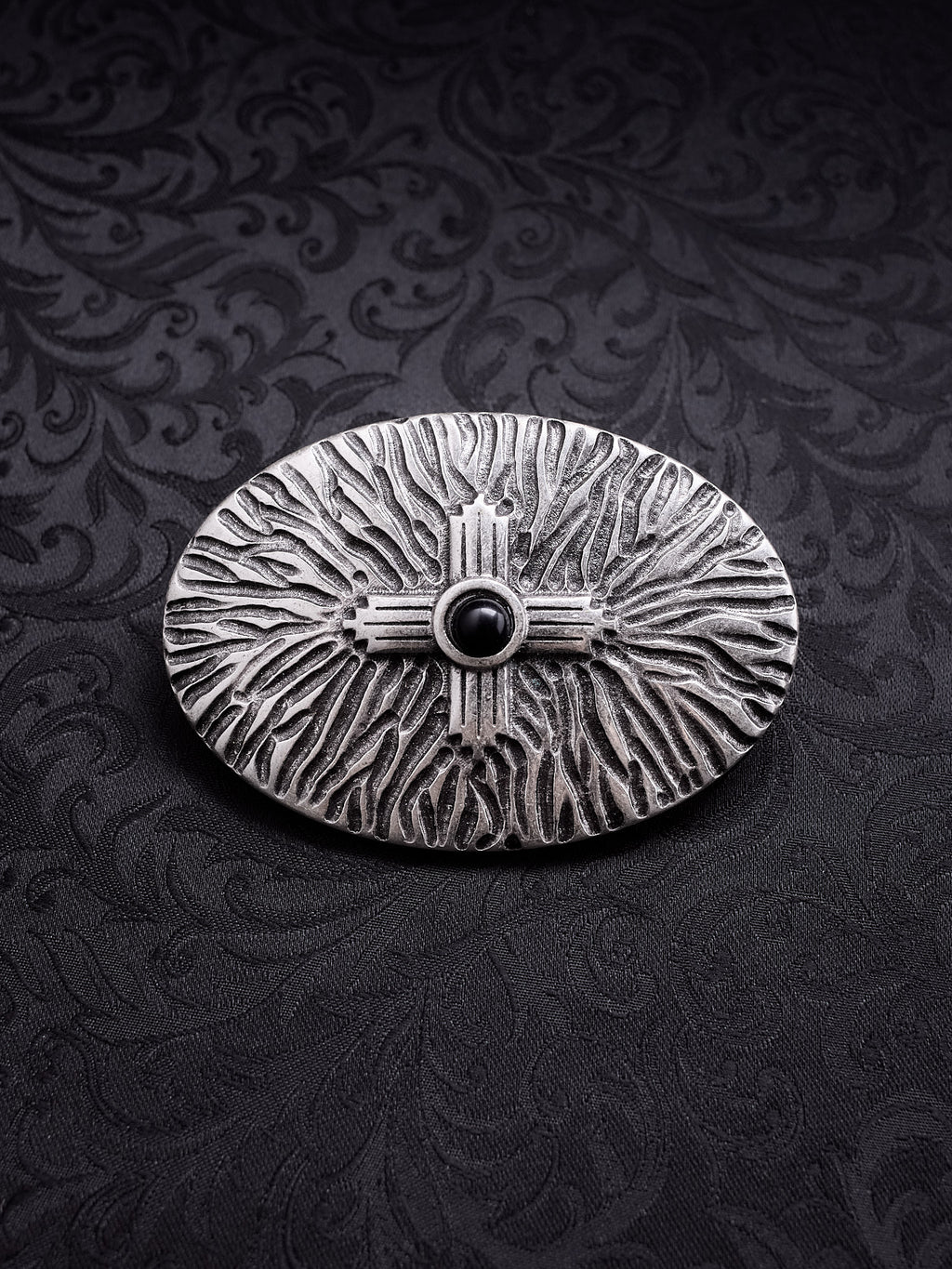 Oval metal belt buckle named Black Halo with a central dark motif and radiating organic textures on a black damask background