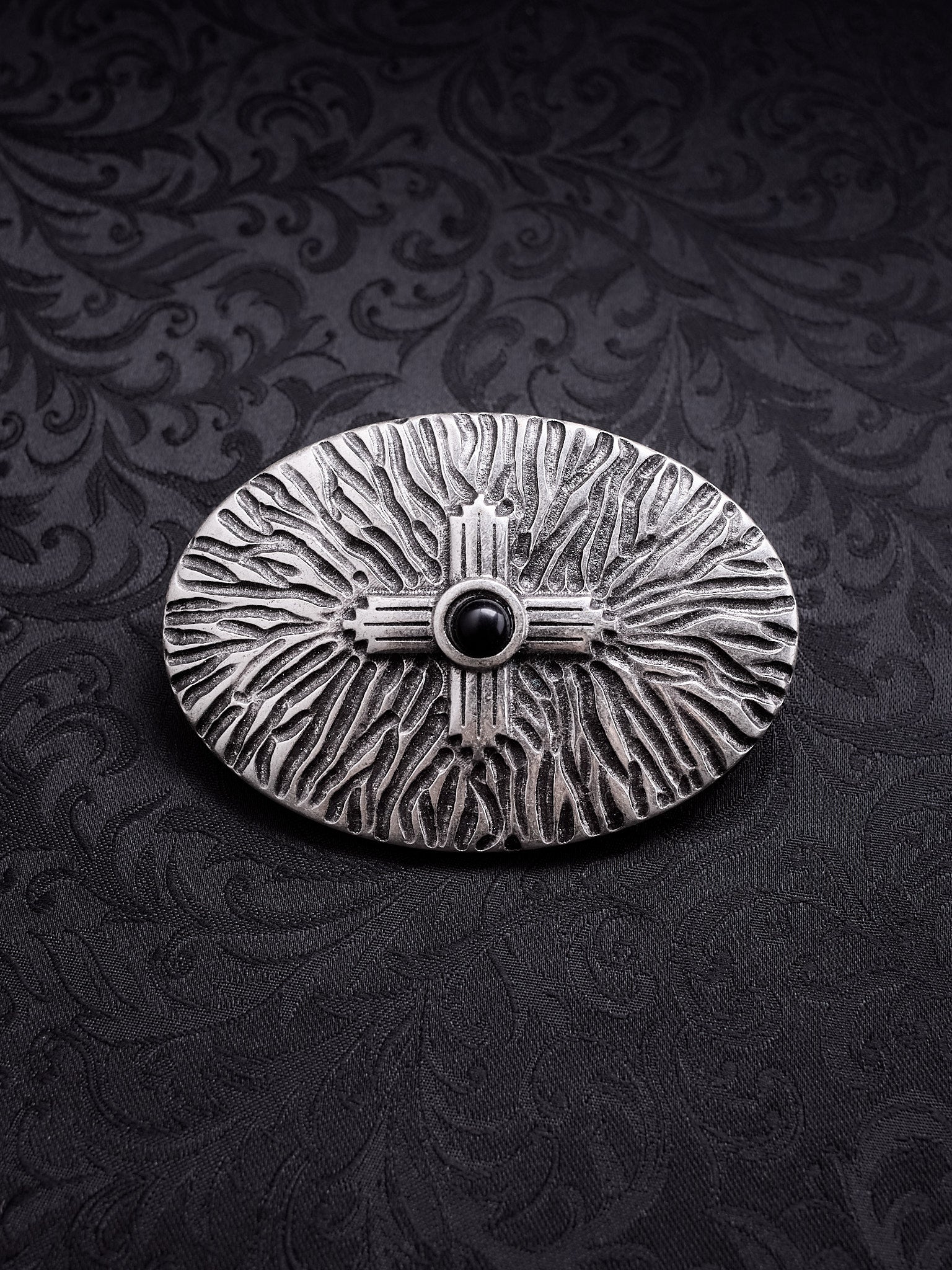 Oval metal belt buckle named Black Halo with a central dark motif and radiating organic textures on a black damask background