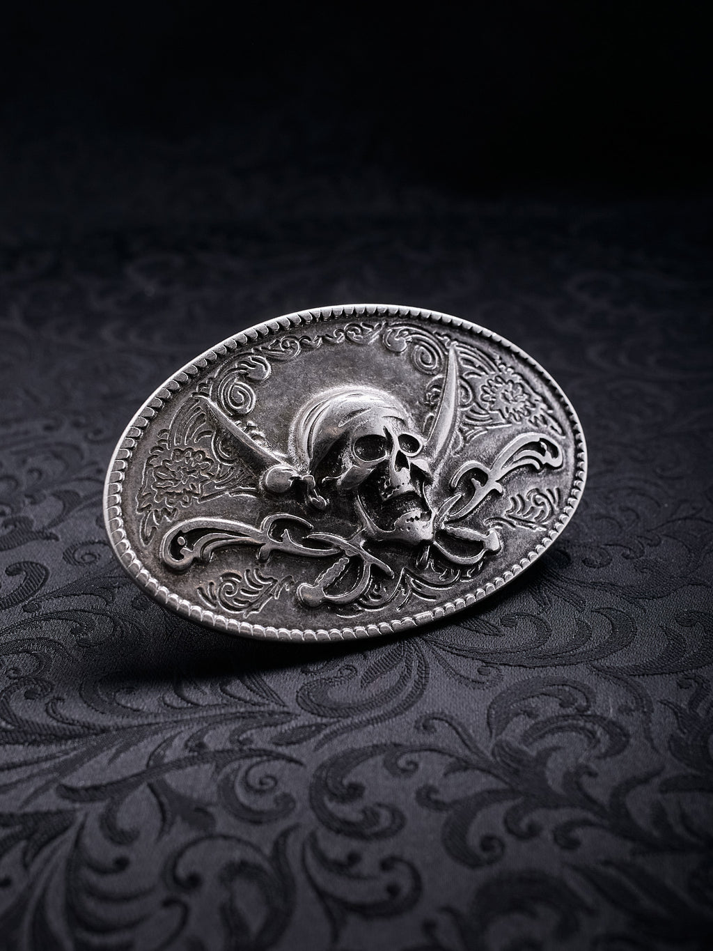 Oval metal belt buckle named Corsair Oath with a skull, crossed sabers, and floral engravings on a black background