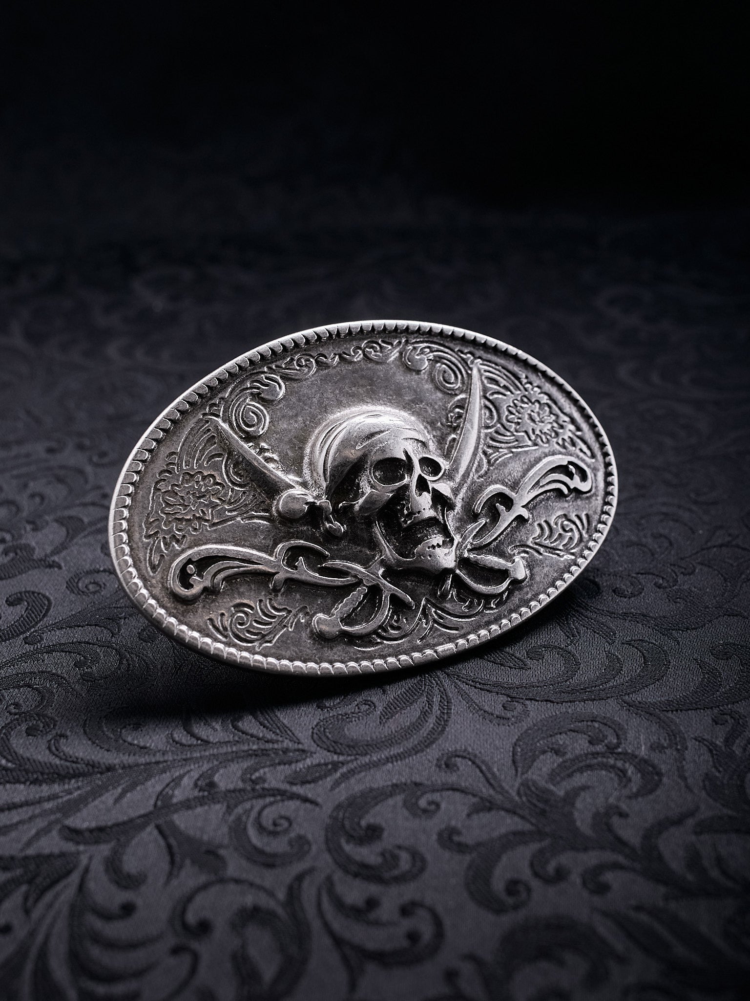Oval metal belt buckle named Corsair Oath with a skull, crossed sabers, and floral engravings on a black background
