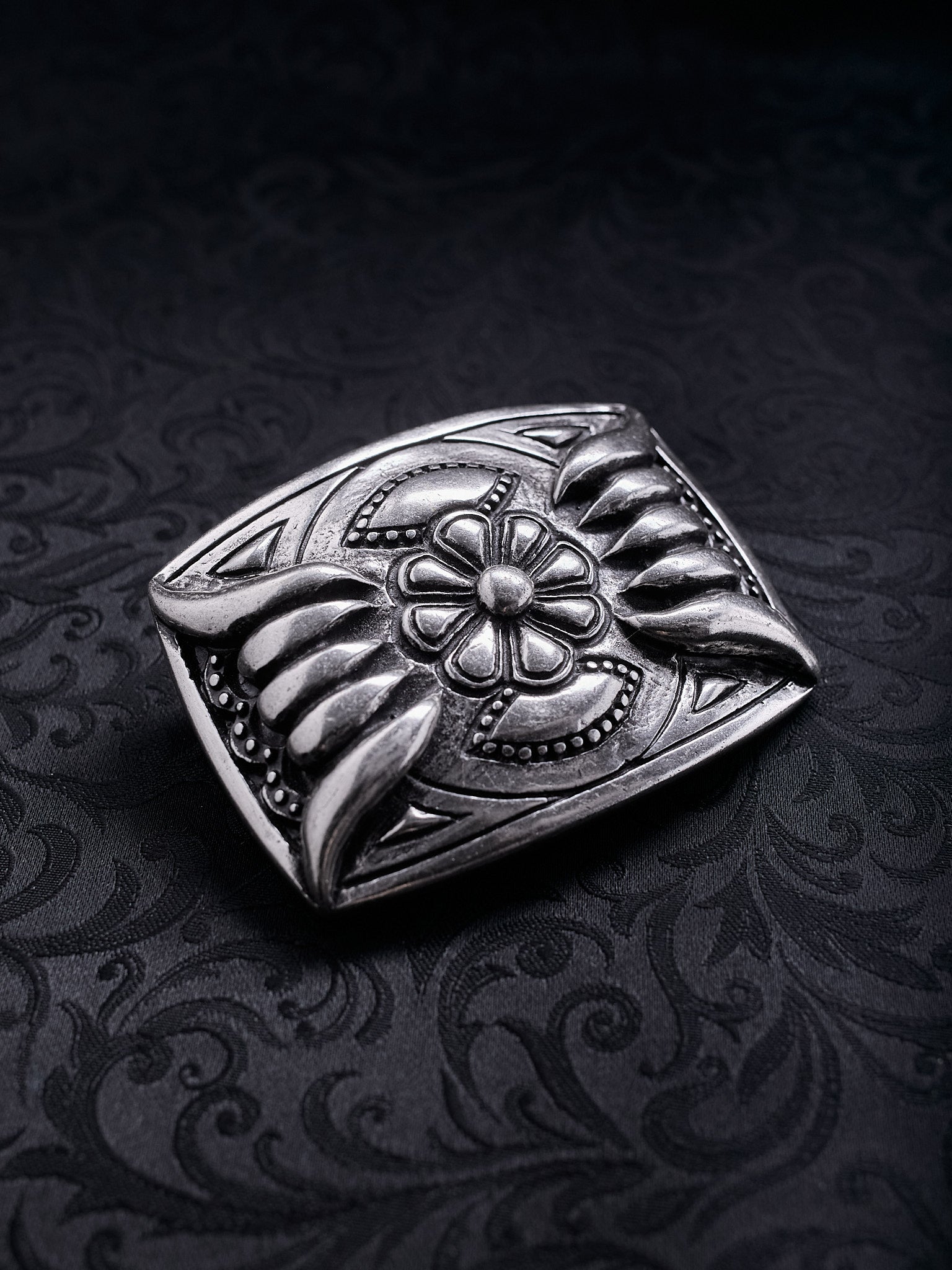 Rectangular metal belt buckle named Iron Bloom with a central flower and claw-like side accents on a dark background