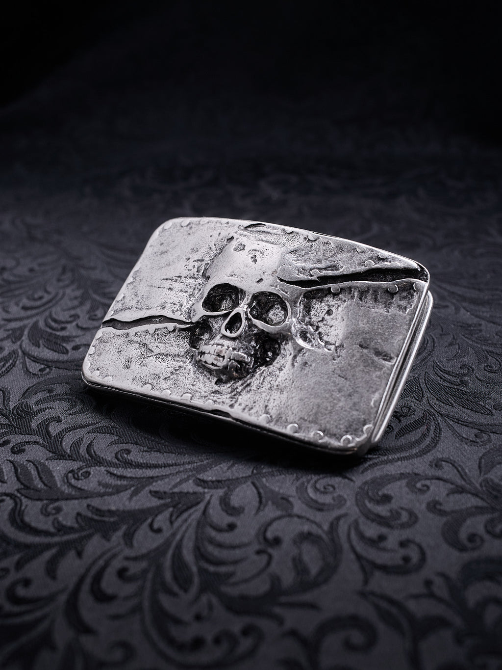 Rectangular metal belt buckle named Memento Fracture with a skull relief and a deep diagonal crack on a dark background