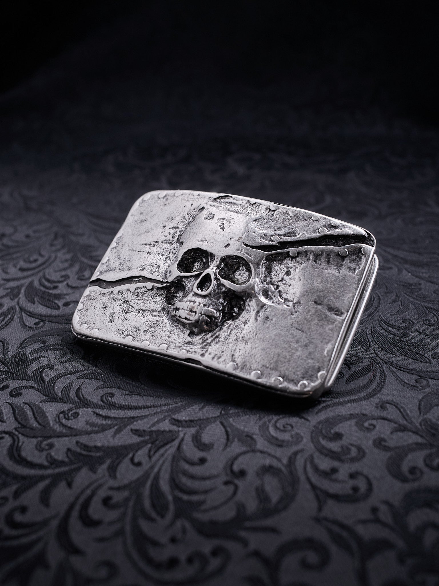 Rectangular metal belt buckle named Memento Fracture with a skull relief and a deep diagonal crack on a dark background