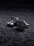 Square metal cufflinks with a complex gothic vine ornament and black oxidized background on a dark damask fabric