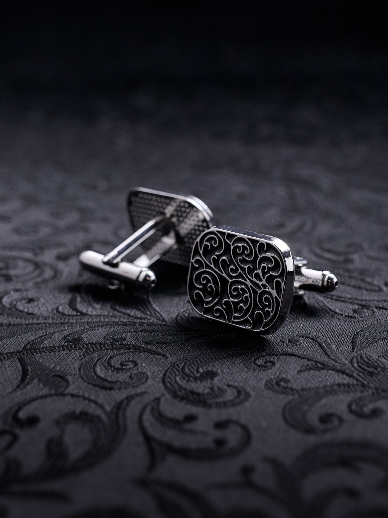 Square metal cufflinks with a complex gothic vine ornament and black oxidized background on a dark damask fabric