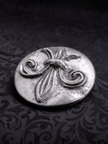 Round metal belt buckle named Sable Fleur with a central Fleur-de-lis relief on a dark background
