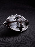 Oval metal belt buckle named Scriptum Obscura with etched calligraphy and a central gothic cross on a dark damask background