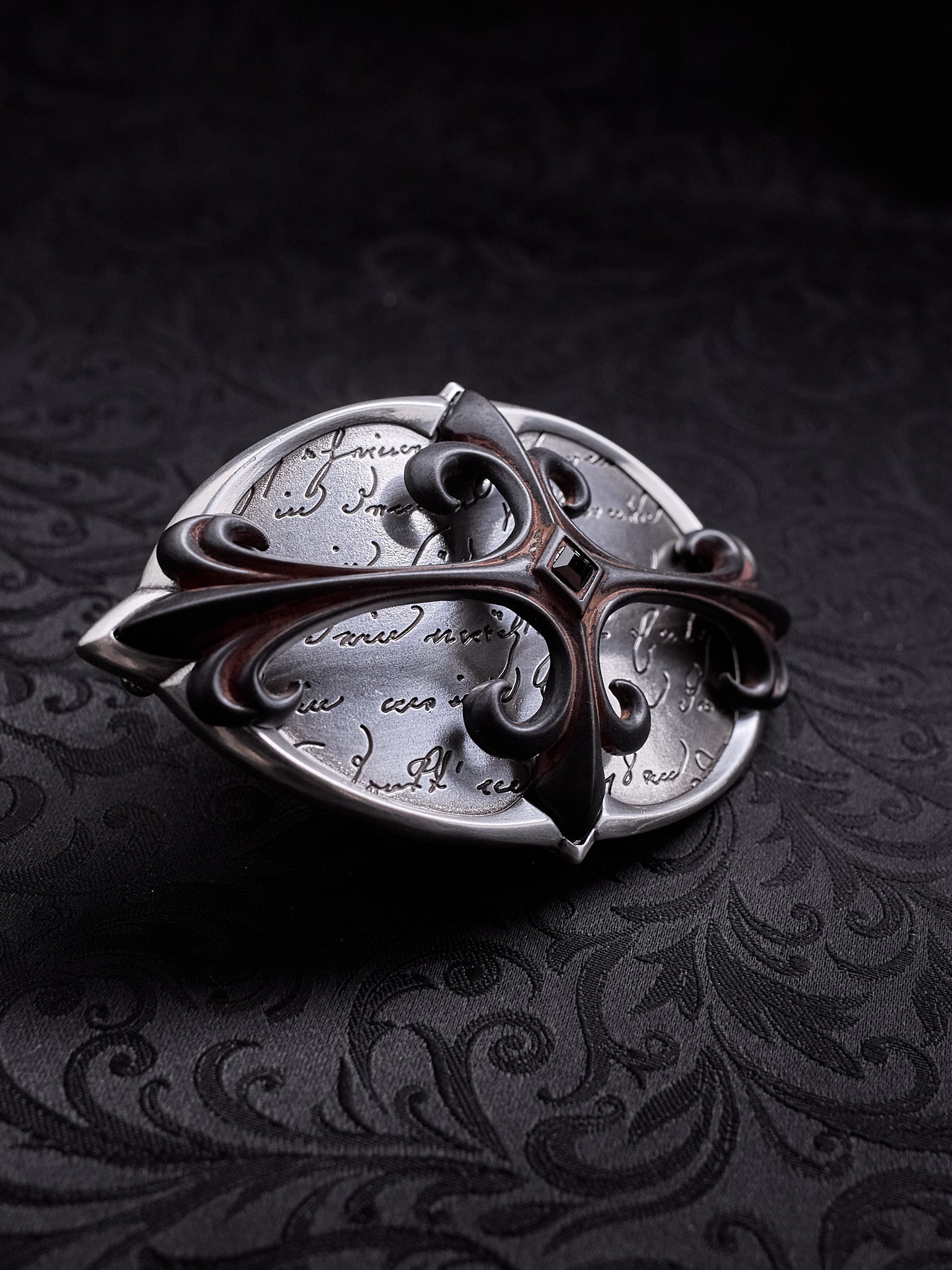 Oval metal belt buckle named Scriptum Obscura with etched calligraphy and a central gothic cross on a dark damask background