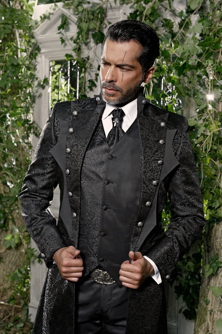 A man wearing a dark gothic-style long coat with ornate silver buttons over a black vest and tie, posed outdoors in front of a white column and green ivy-covered walls.