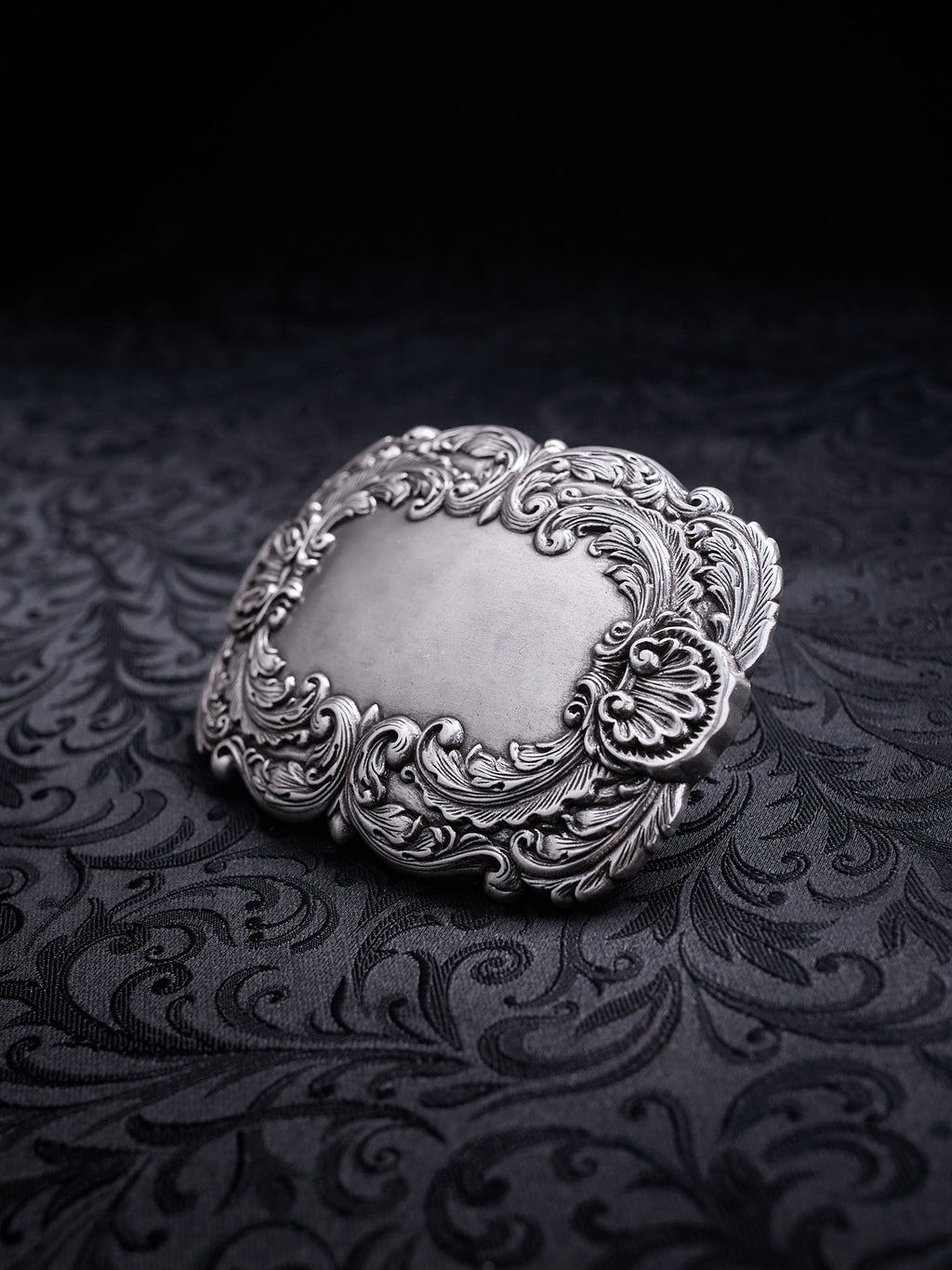 Ornate metal belt buckle named Velour Nocturne with a decorative floral baroque frame on a black background