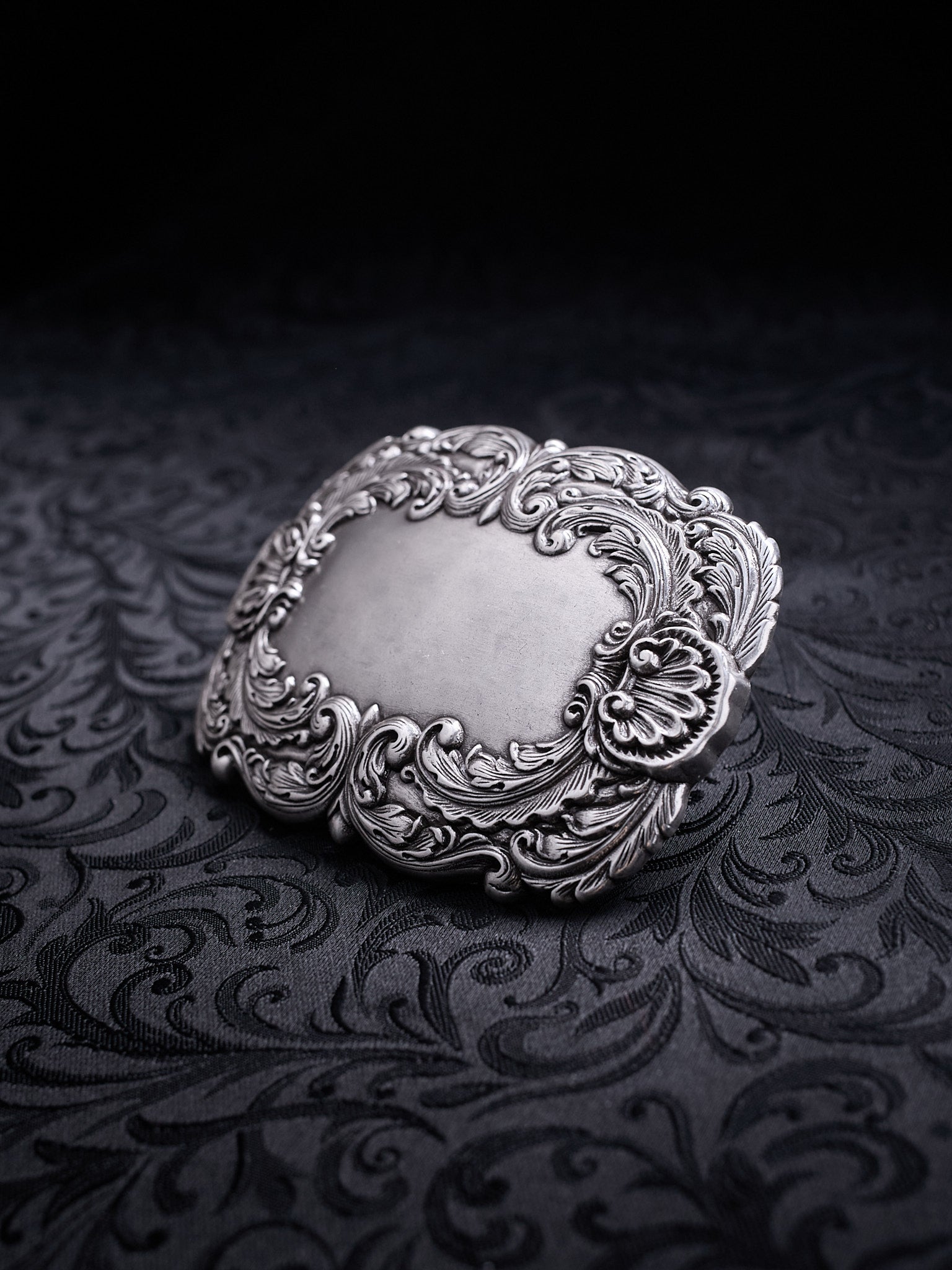 Ornate metal belt buckle named Velour Nocturne with a decorative floral baroque frame on a black background